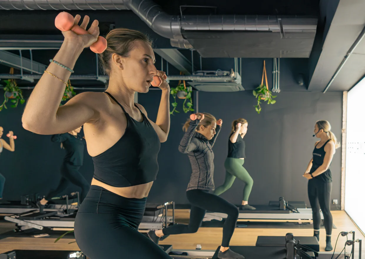 Clients in a Reformcore class performing weighted lunges on the Reformer, combining strength training, stability, and dynamic movement in a premium Pilates studio.