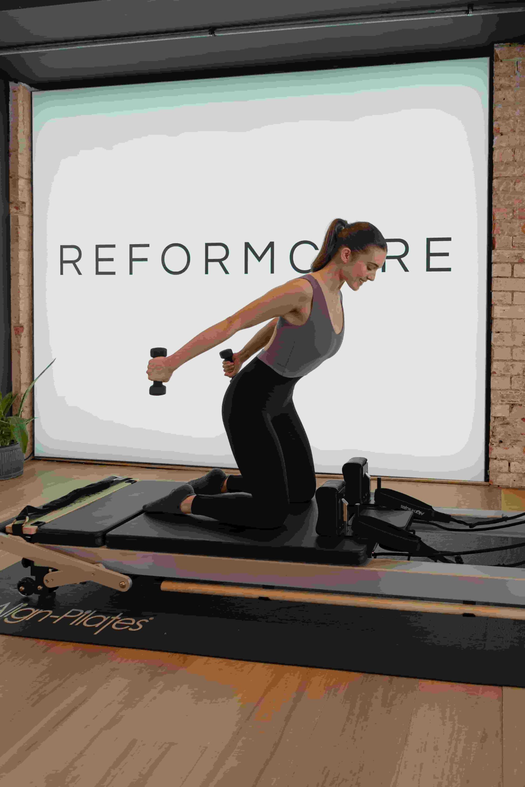 Client kneeling on the Reformer during a Reformcore Pilates class, performing tricep kickbacks with dumbbells to build upper body strength and control in a refined studio setting.