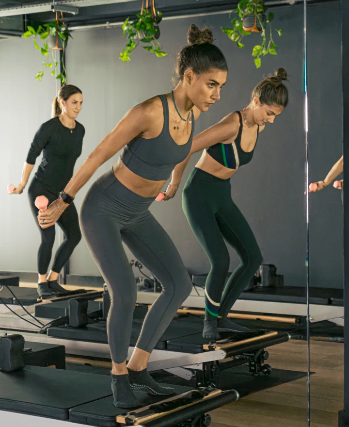 Group of women in a Reformcore Reformer Pilates class performing a dynamic arm and core sequence with light dumbbells, combining resistance and balance training for full-body strength in a sleek, modern studio.