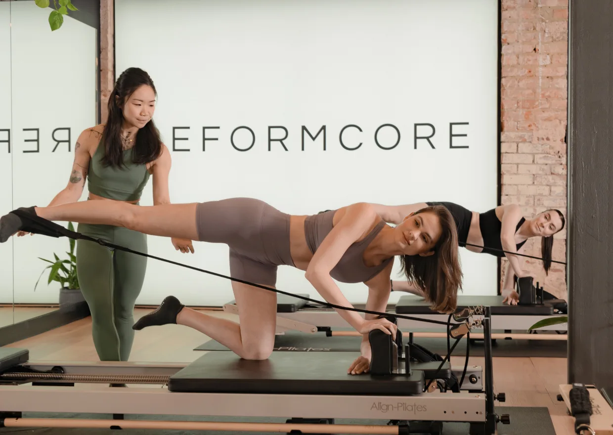 Reformcore instructor supporting a client through a side-lying leg lift on the Reformer, focusing on glute strength, core engagement, and proper alignment in a premium Pilates class.